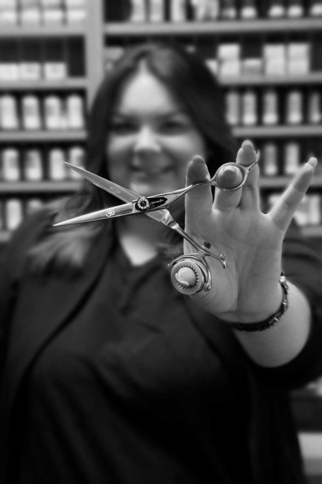 Hairdresser holding scissors, smiling in salon with shelves of hair products in the background.